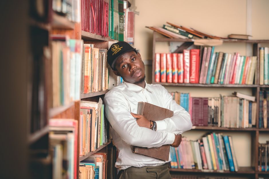 Young man in a Ghanaian library holding a book, surrounded by bookshelves and literature.
